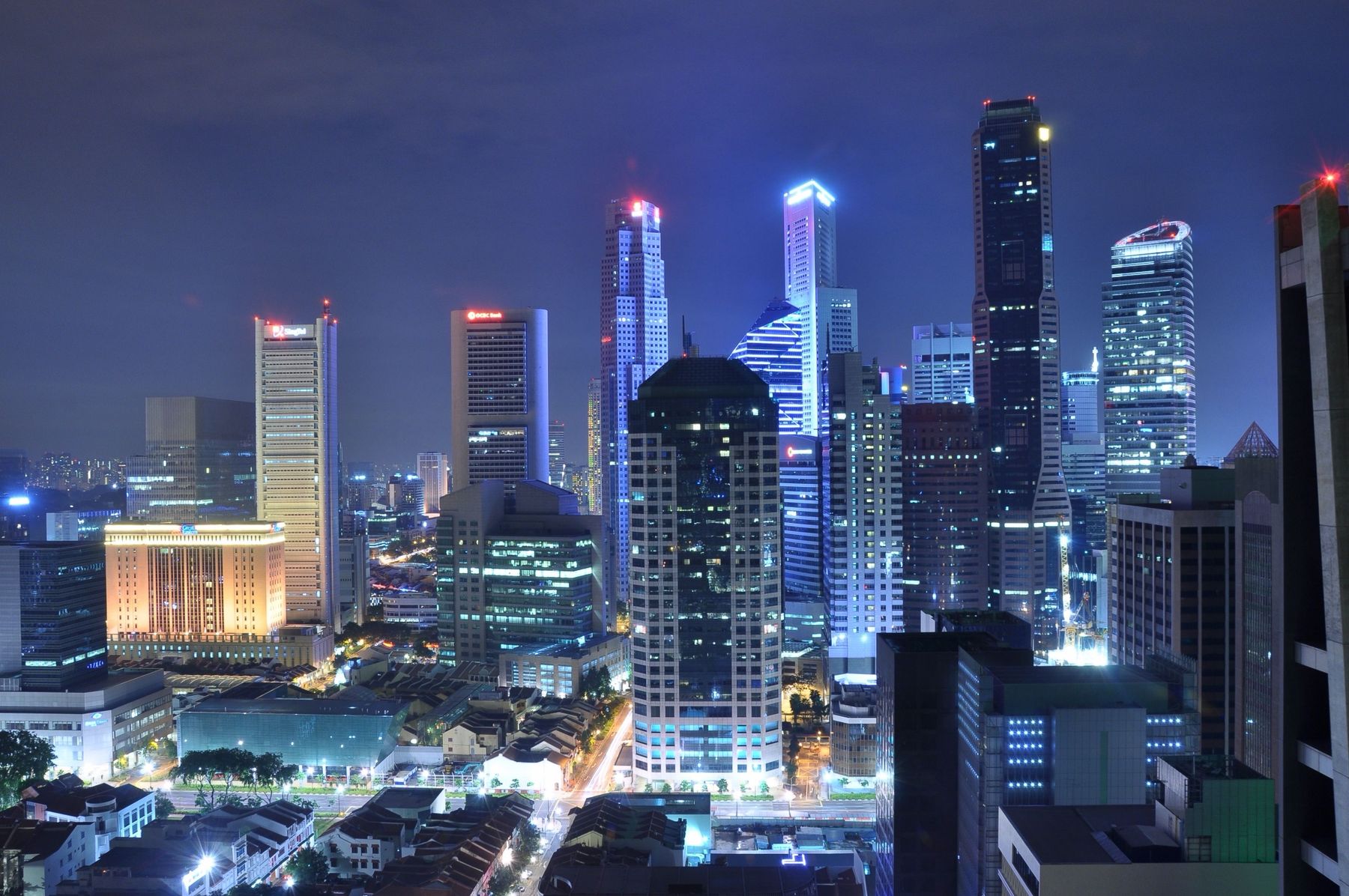 Skyscrapers lit blue at night above old shophouse roofs